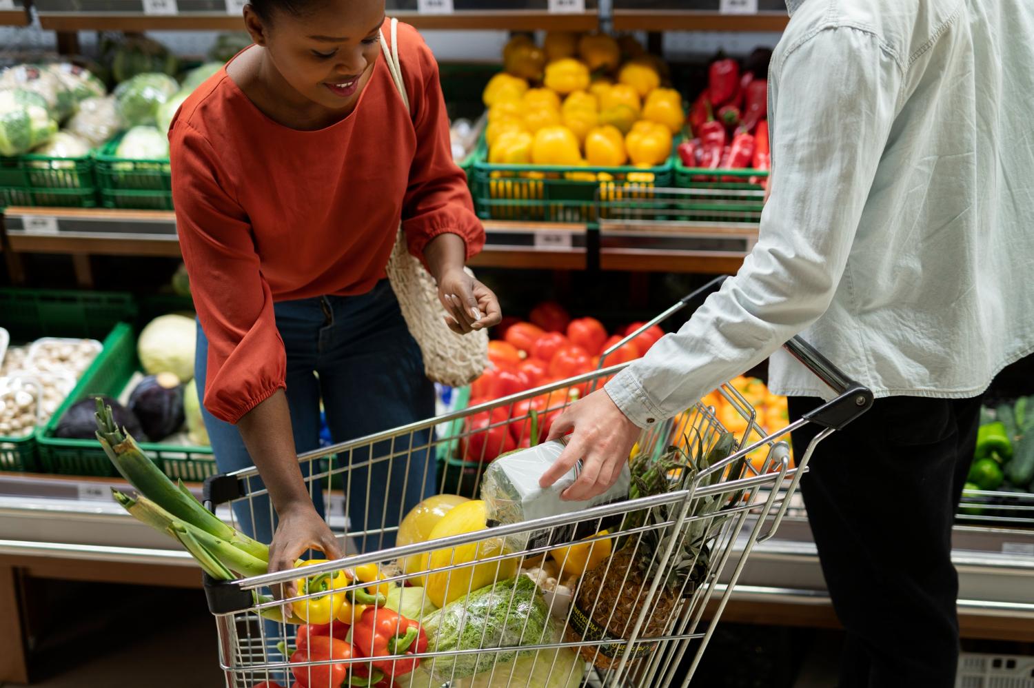 Uma mulher está fazendo compras em um supermercado com um homem. Ela tem uma cesta cheia de frutas e legumes, incluindo cenoura e brócolis. O homem está segurando a cesta enquanto ela olha seus itens. Eles estão cercados por outras pessoas na loja, algumas das quais também carregam sacolas ou carrinhos de compras.