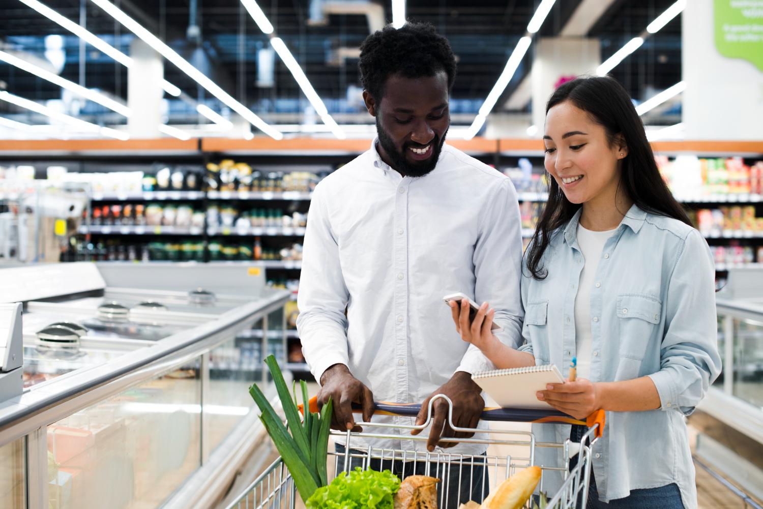 Num supermercado bem iluminado, um homem e uma mulher fazem compras juntos. O homem veste uma camisa branca com as mangas arregaçadas até os cotovelos, enquanto a mulher veste uma jaqueta jeans azul por cima de um top de cor clara. Ambos têm carrinhos cheios de mantimentos, incluindo produtos frescos como cenoura e brócolis. Seus sorrisos sugerem que estão gostando da experiência de compra.